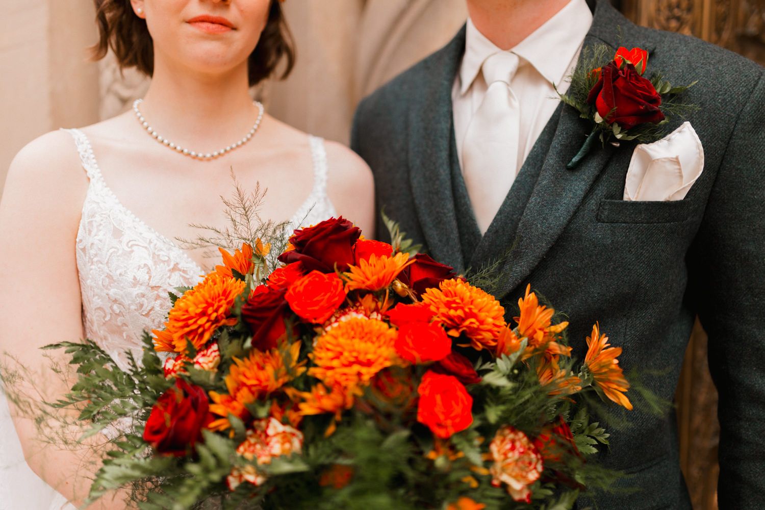 A couple in wedding attire poses together with vibrant red and orange floral bouquets.