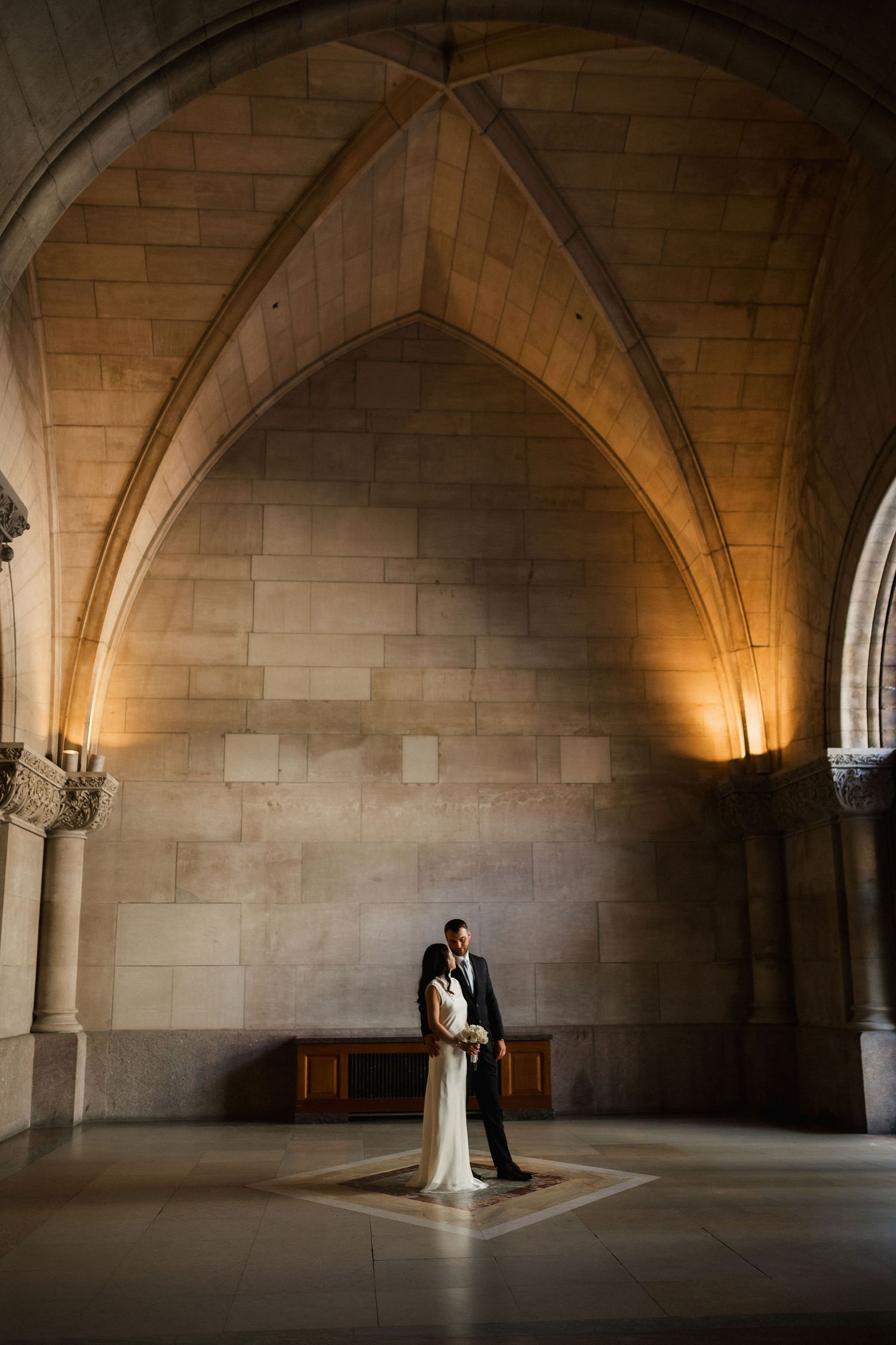 Bride and groom embracing under Gothic stone arches in a grand cathedral hall, warmly lit for a romantic wedding portrait.