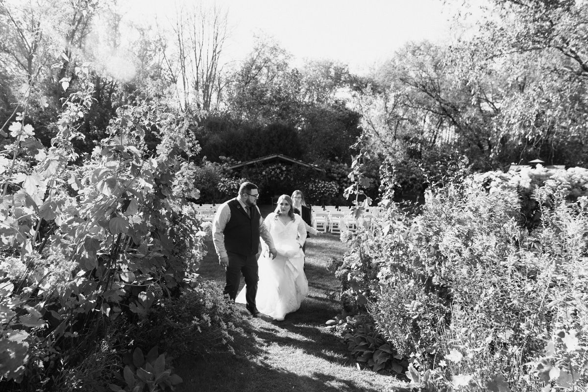 A black and white photograph of a couple walking through a lush garden path surrounded by flowering bushes and trees.