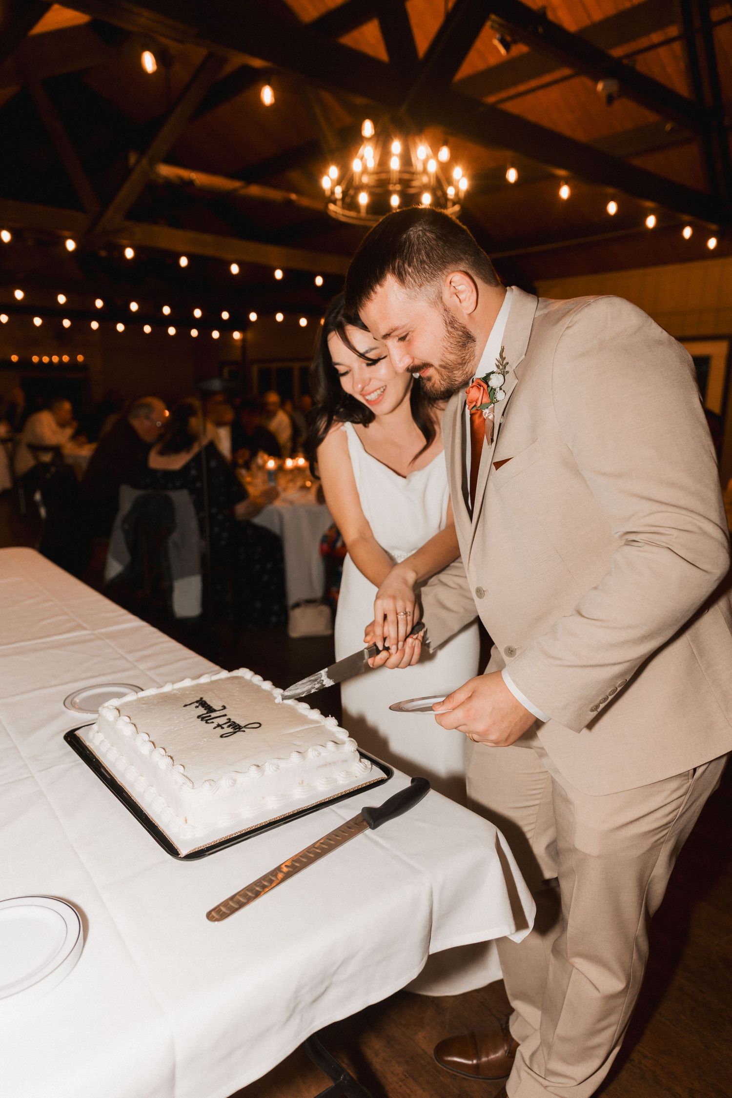 A couple in wedding attire cuts their white wedding cake together at an indoor reception with string lights overhead.