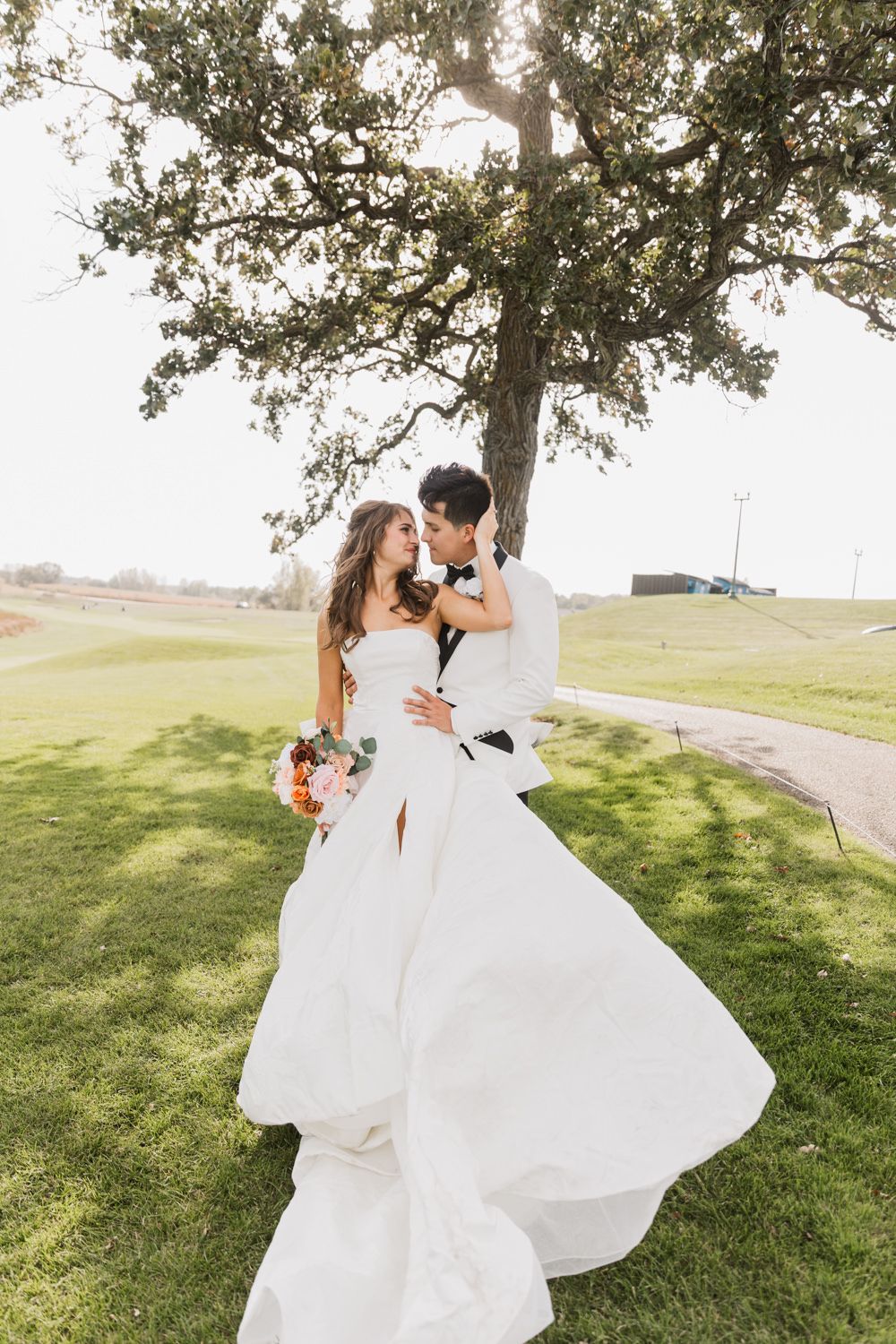 A romantic wedding couple dances together on a grassy hillside as the bride's white dress flows elegantly in the breeze.
