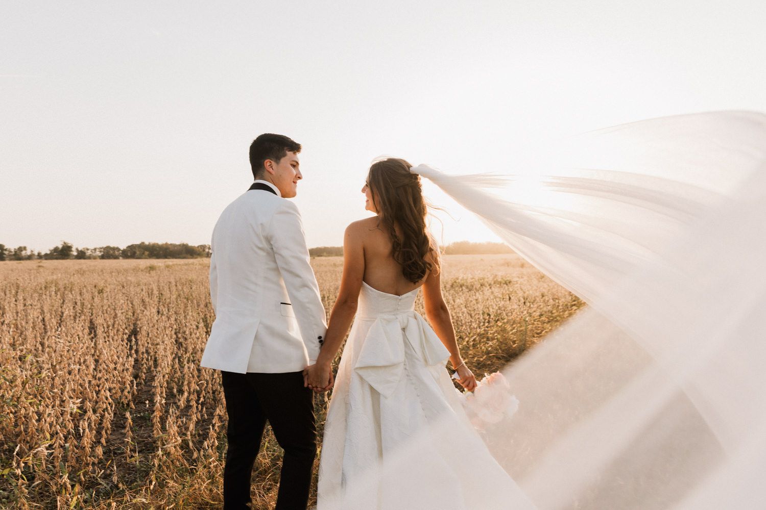 A couple stands together in a golden wheat field as a white veil flows dramatically in the wind.