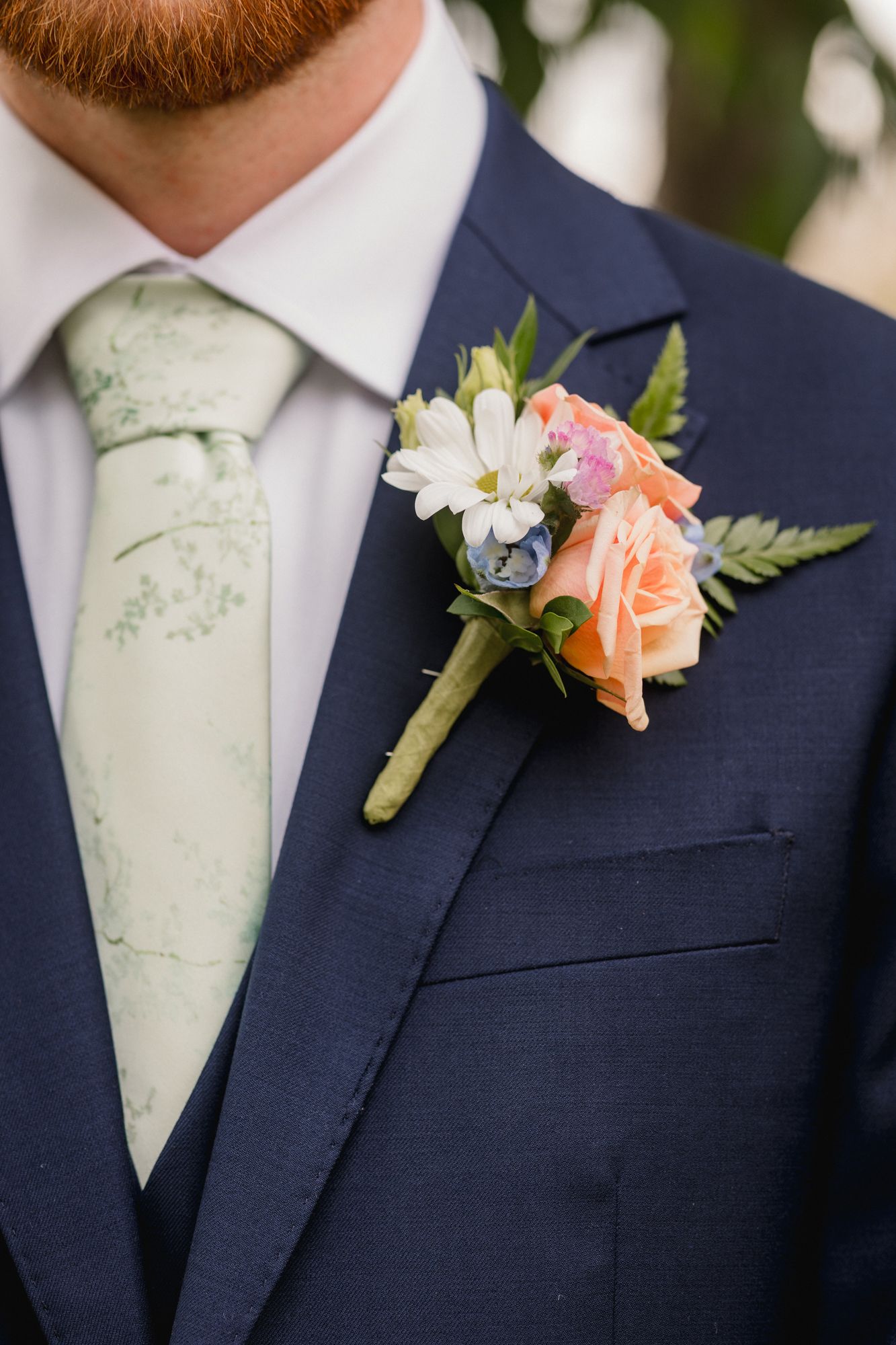 Groom in navy suit with light green floral tie and peach rose boutonniere with white daisies and greenery.