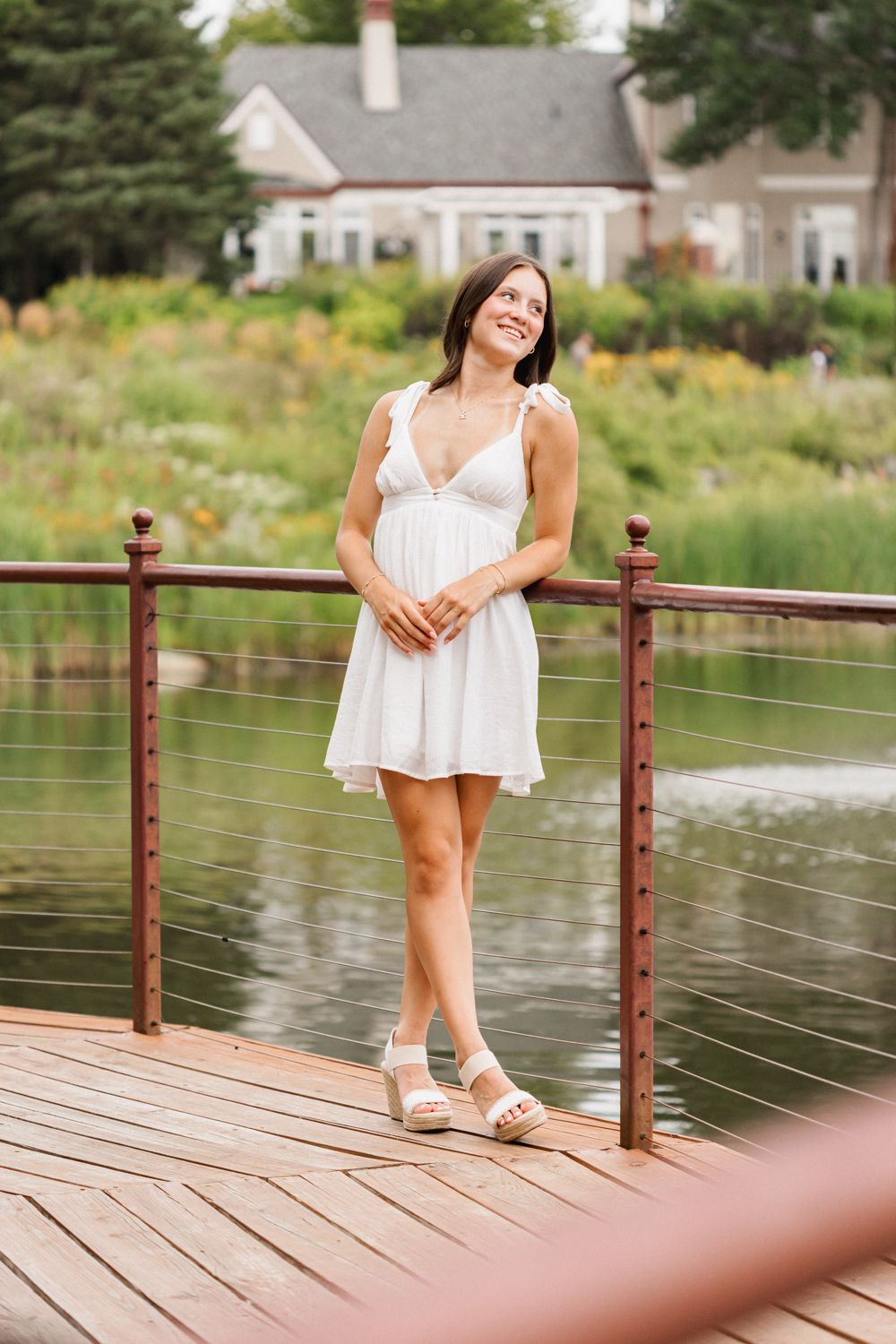 A person in a white summer dress poses on a wooden deck overlooking a pond with a house in the background.