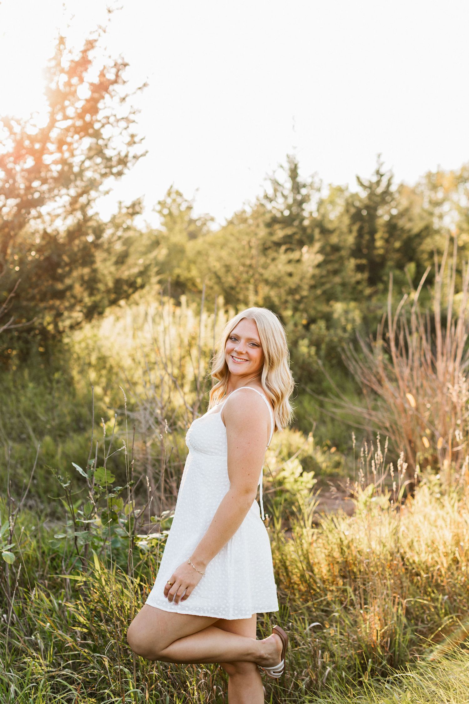 A person in a white top poses outdoors in tall grass during golden hour sunset lighting.