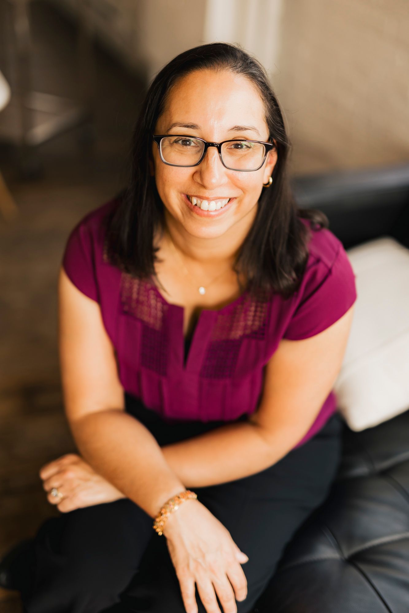 Professional headshot of person in magenta top and glasses sitting casually indoors.