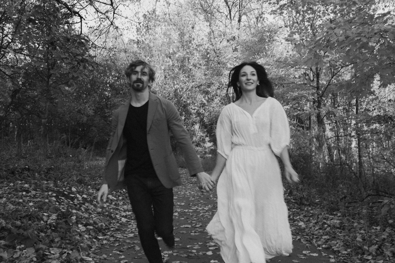 A couple walks together through a forest path with the woman wearing a flowing white dress and man in dark attire.
