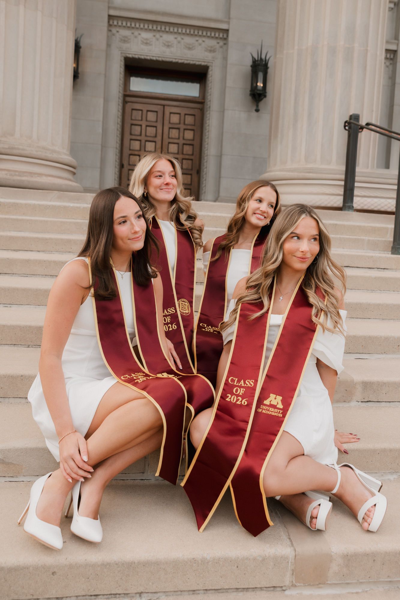 Four female graduates in white dresses and maroon Class of 2026 sashes pose on university steps.