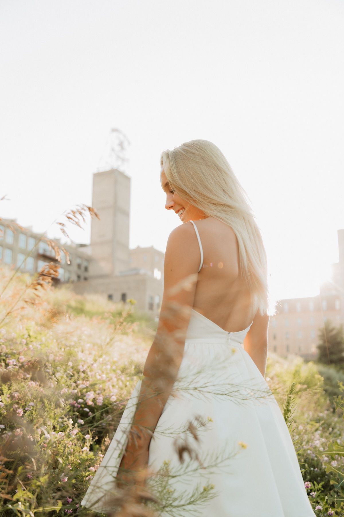 Blonde woman in white backless dress smiling among wildflowers with sunlit urban backdrop.