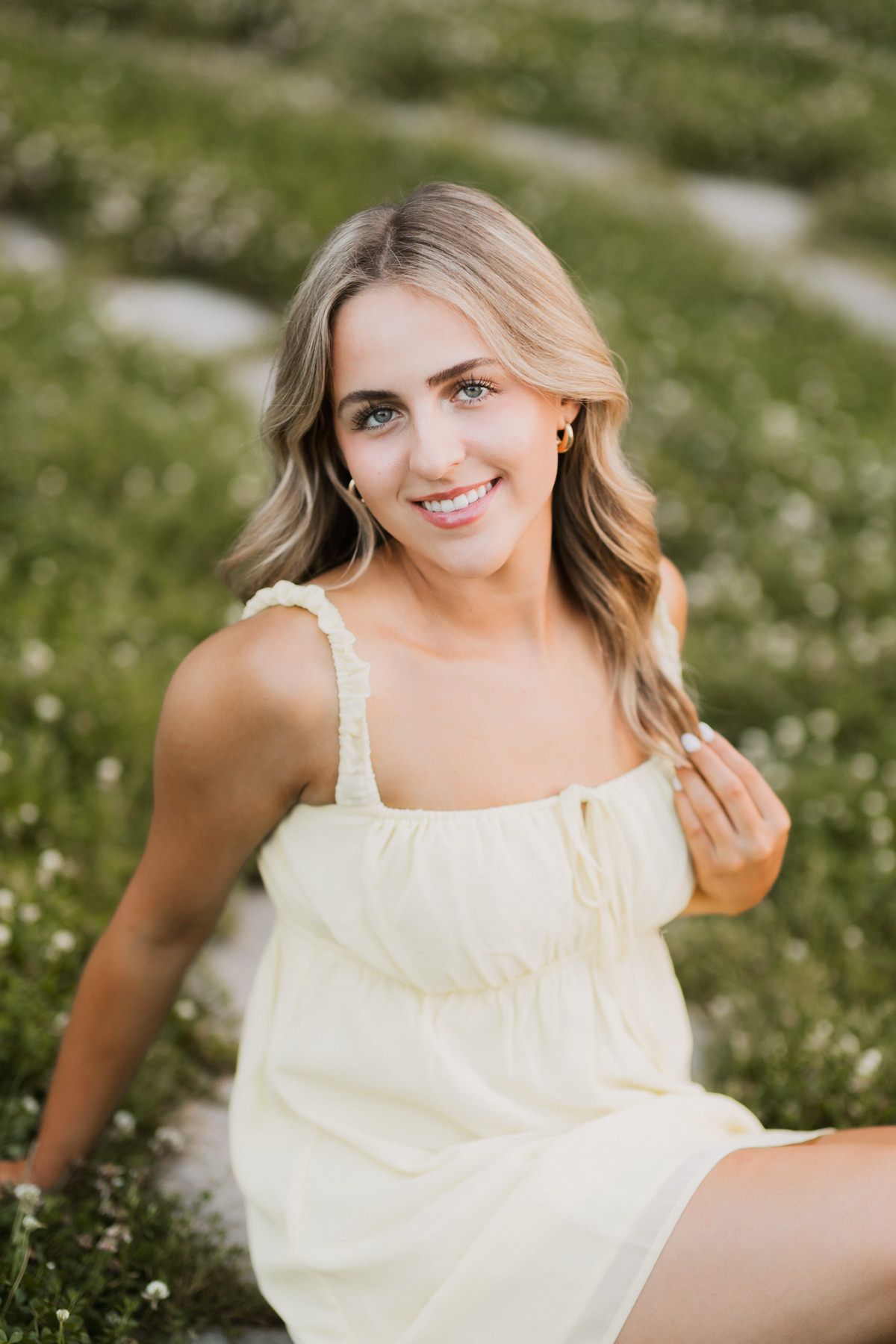 Smiling blonde woman in yellow sundress sitting in a field of white wildflowers outdoors.