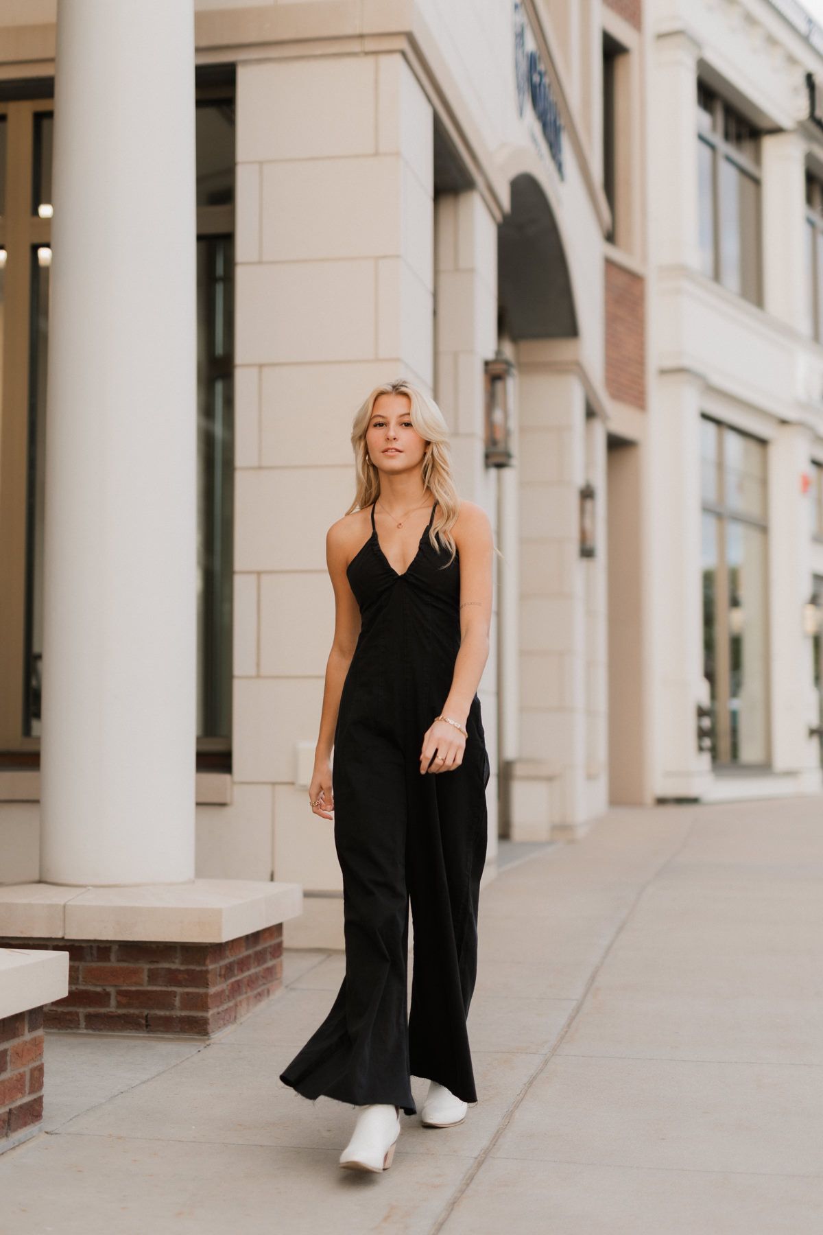 Blonde woman in black halter jumpsuit walking on urban sidewalk near stone columns and storefronts.