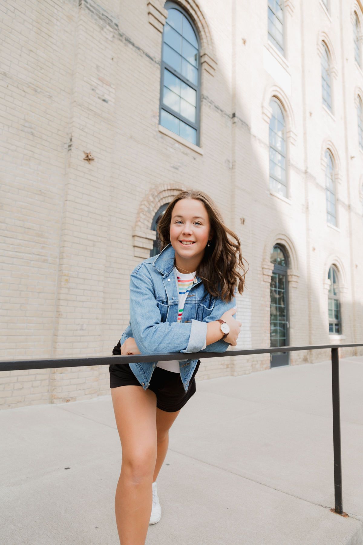 Smiling teen girl in denim jacket and black shorts leaning on railing outside brick building.