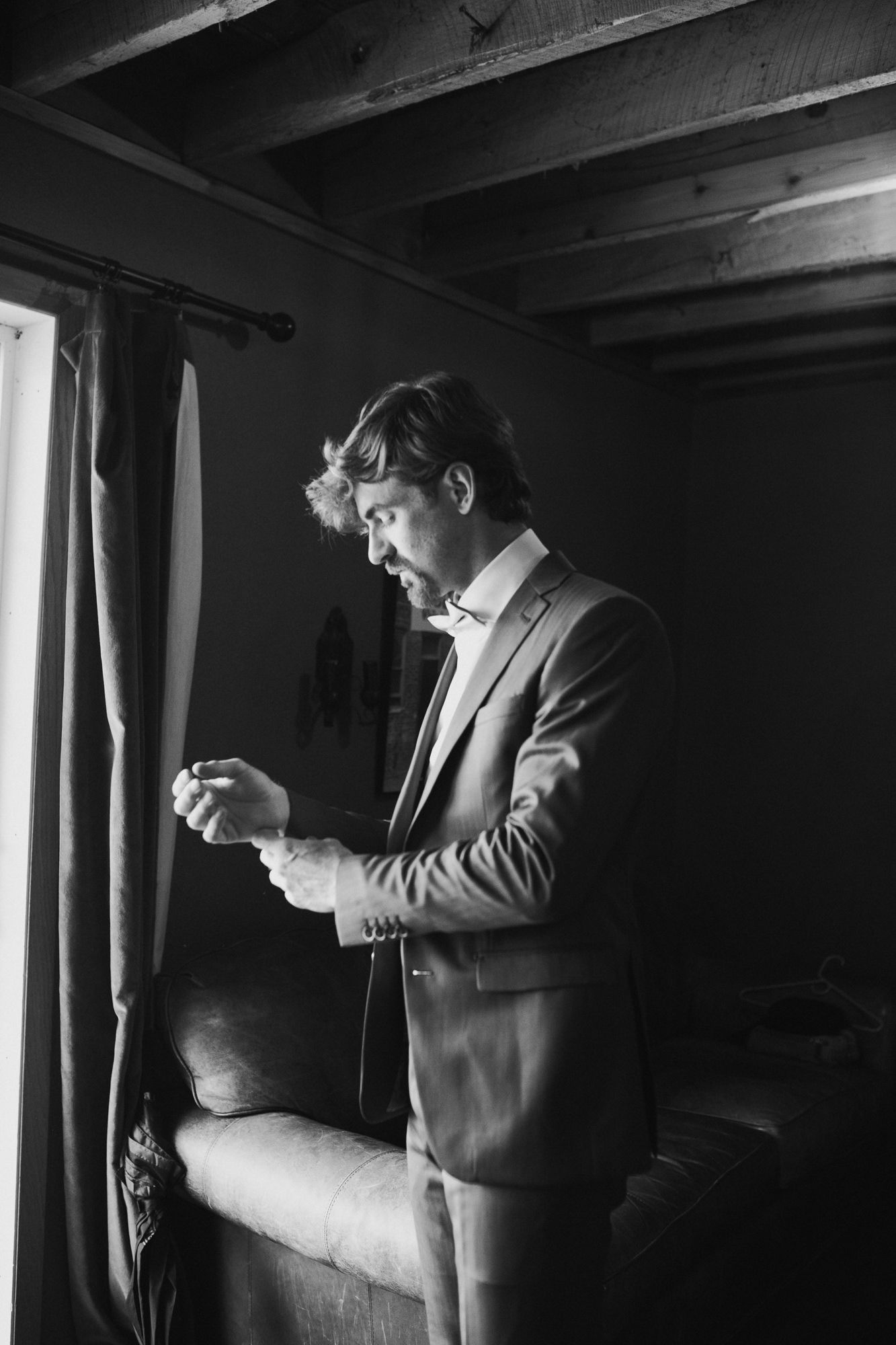 Black and white photo of a well-dressed man in a suit adjusting his cufflinks by a window in a dimly lit room.