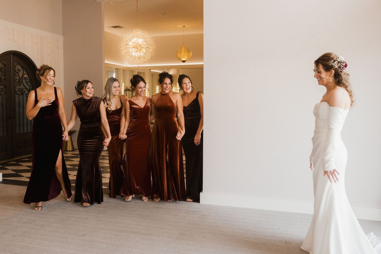 Bridesmaids in burgundy velvet dresses smile as they see the bride in an elegant white off-the-shoulder gown during a first look.