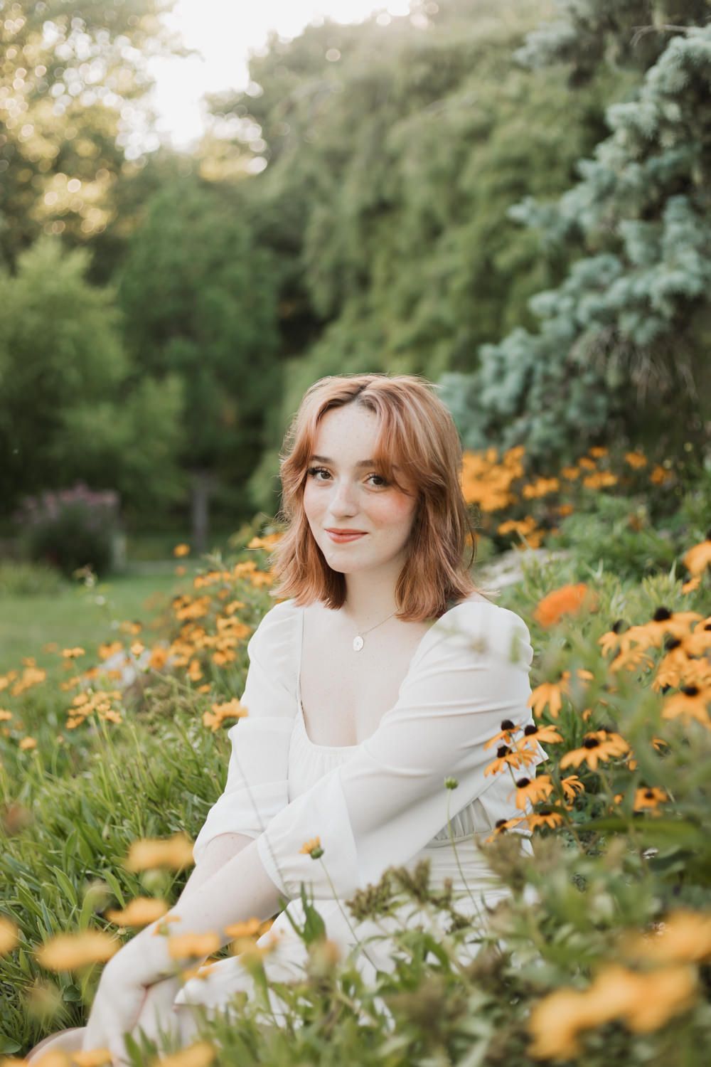 A person in a white dress poses among blooming yellow wildflowers and evergreen trees during golden hour.