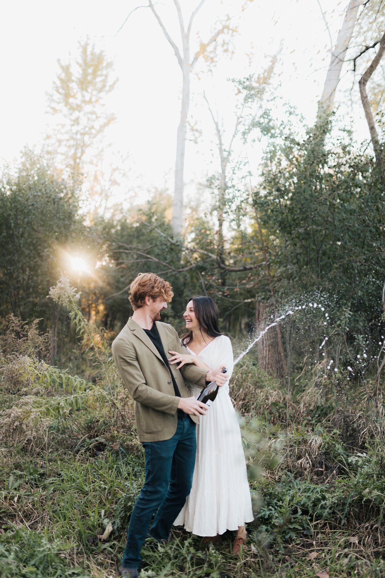 A couple celebrates with champagne spray in a scenic outdoor setting at sunset.