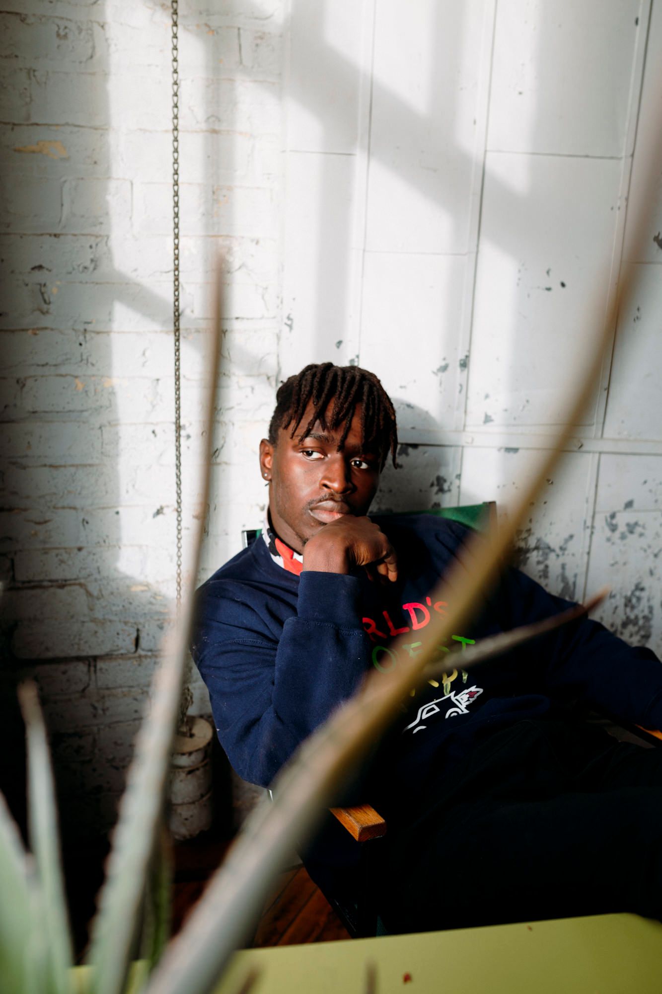 Young man in navy sweatshirt sits thoughtfully against white brick wall, framed by green plants in natural light.