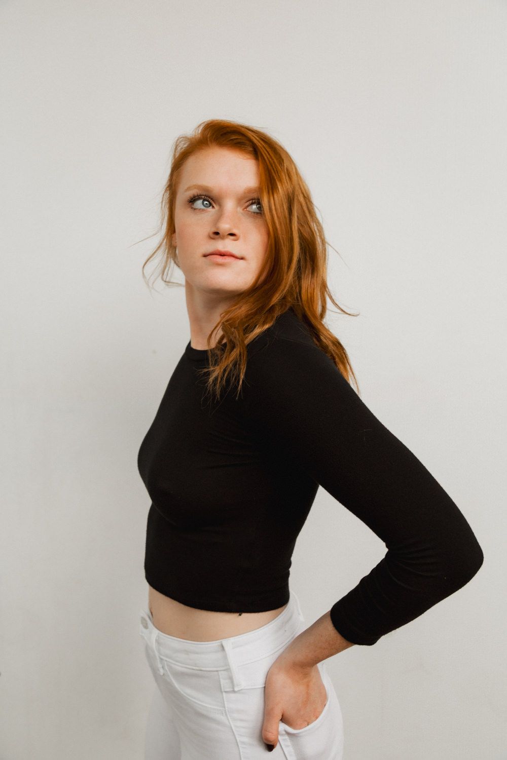 Young woman with red hair wearing a black crop top and white jeans posing against a white background.