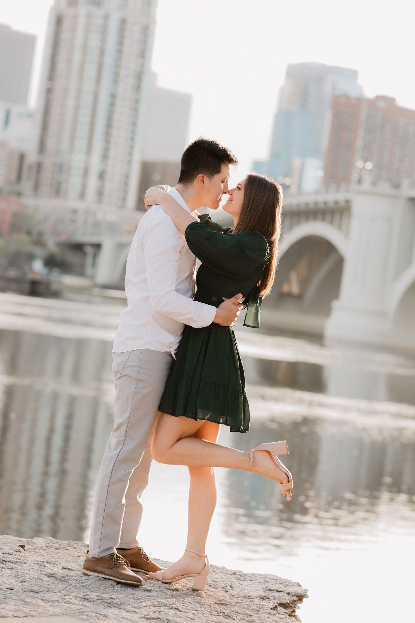 Couple sharing a romantic kiss by a river with a city bridge and skyline in the background on a sunny day.