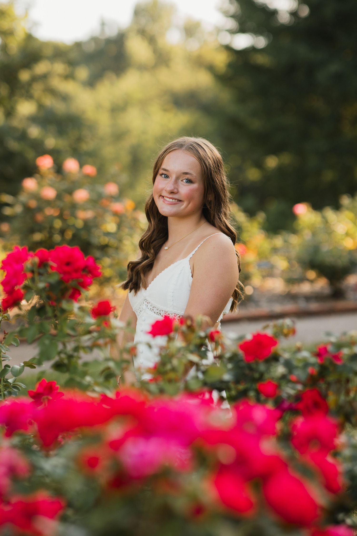 Young woman in white dress smiling among vibrant red roses in a sunlit garden.