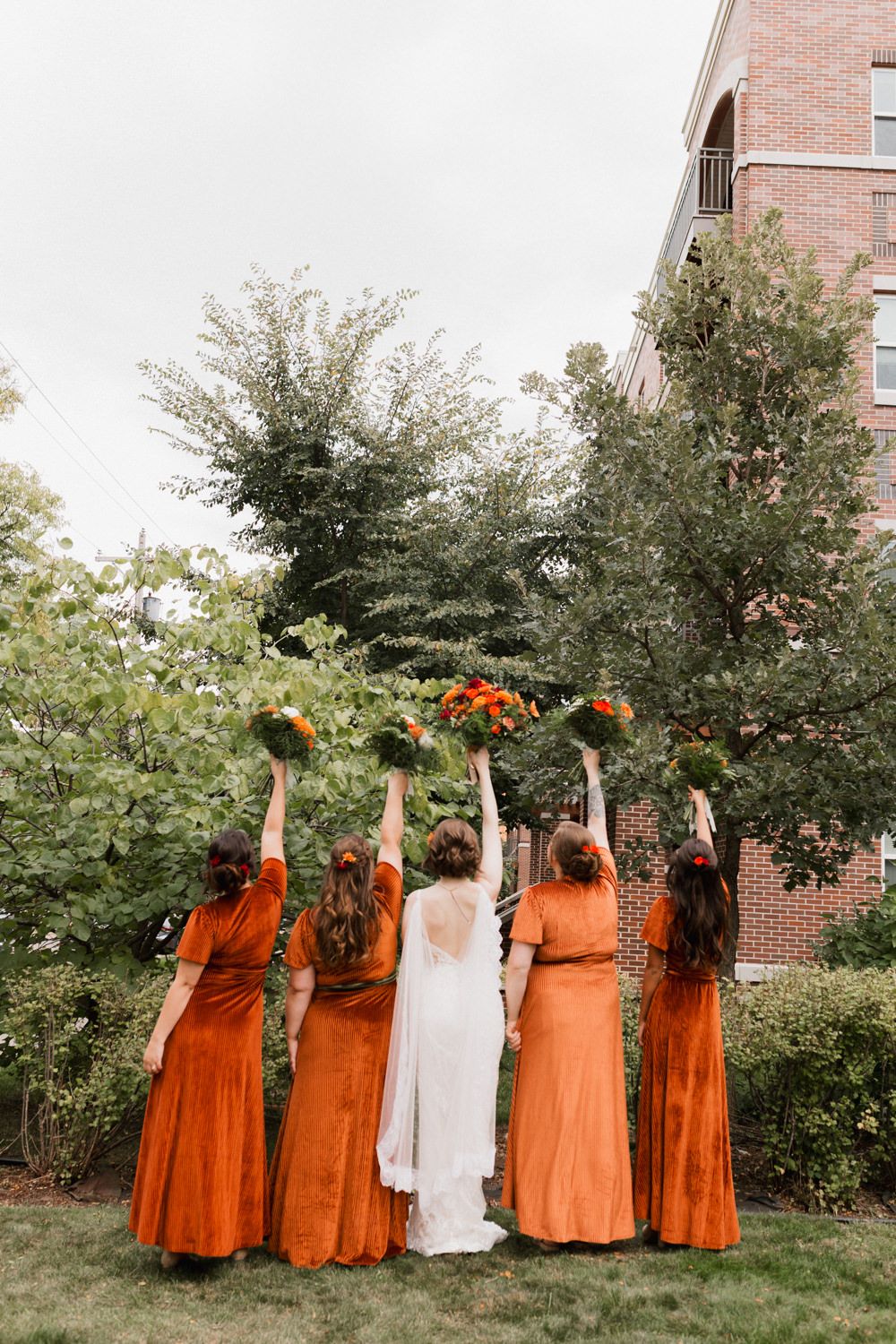 Wedding party poses outdoors in coordinated burnt orange bridesmaid dresses against brick building and greenery.