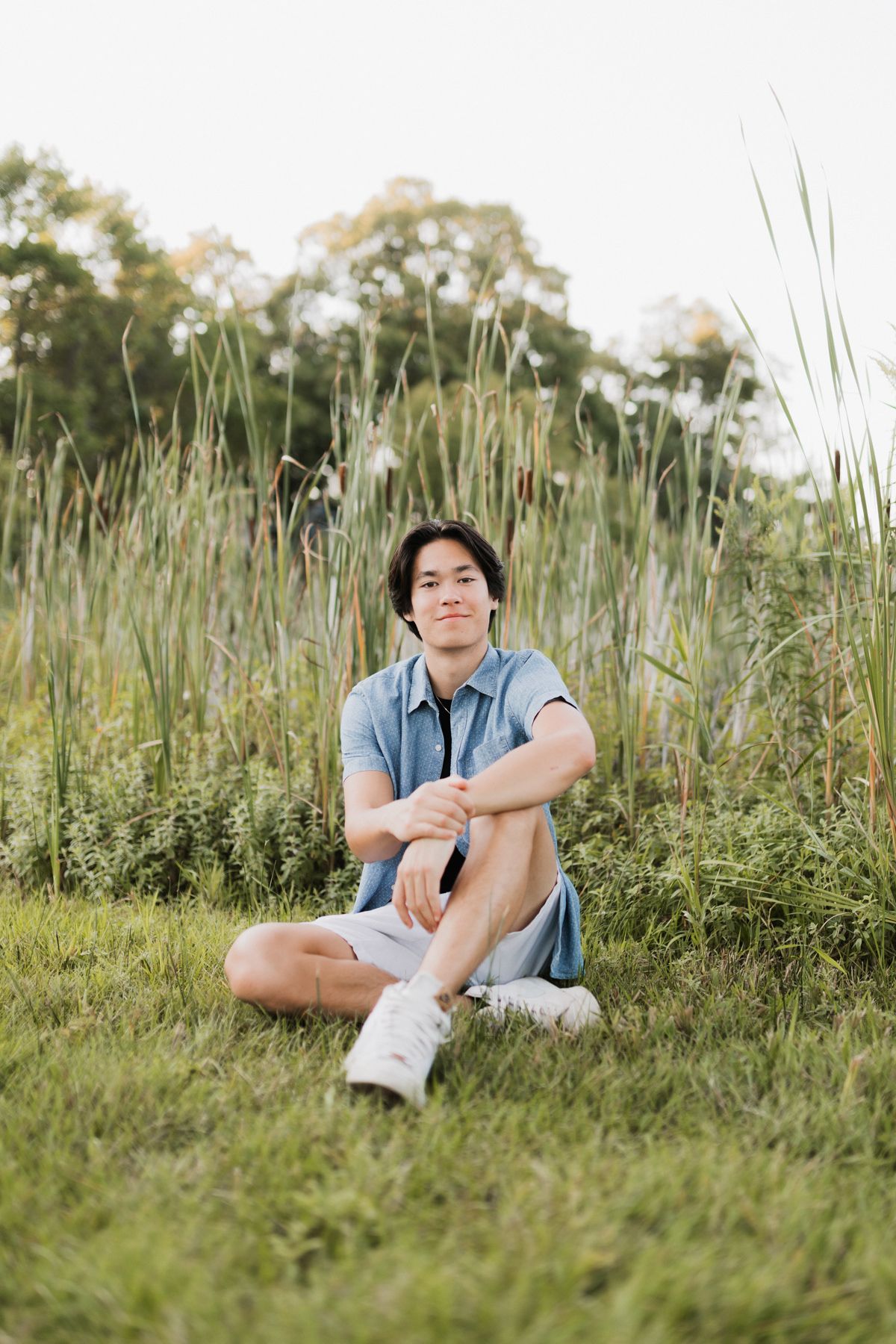 Young man sitting cross-legged on grass near tall cattails, wearing blue shirt and white shorts, smiling at camera.