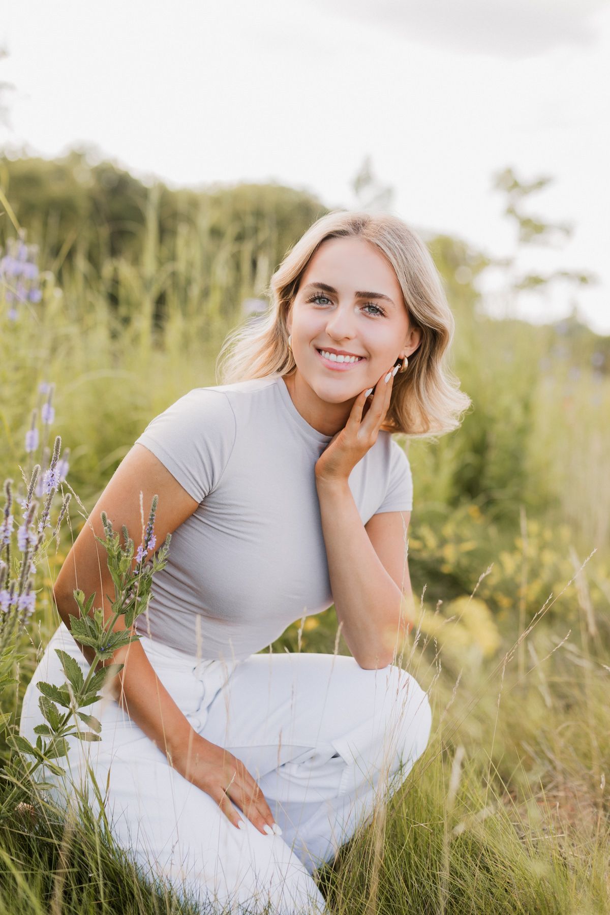 Smiling blonde woman in grey top and white pants poses in wildflower meadow.