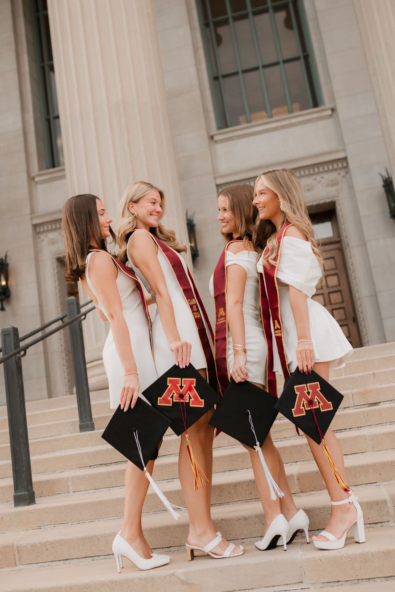 Four female University of Minnesota graduates in white dresses holding caps on stone steps.