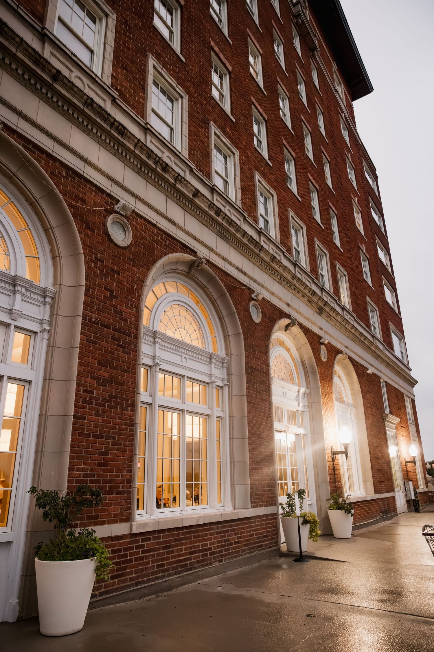 Historic red brick building with arched windows glowing warmly at dusk, featuring elegant white trim and potted plants.