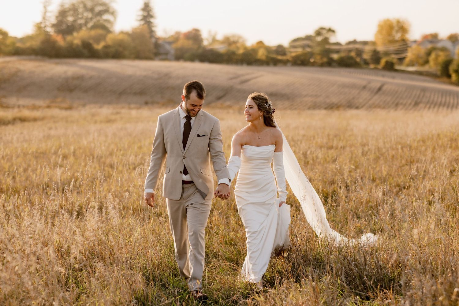 Bride and groom walk hand-in-hand through golden field at sunset, she in white gown, he in gray suit.