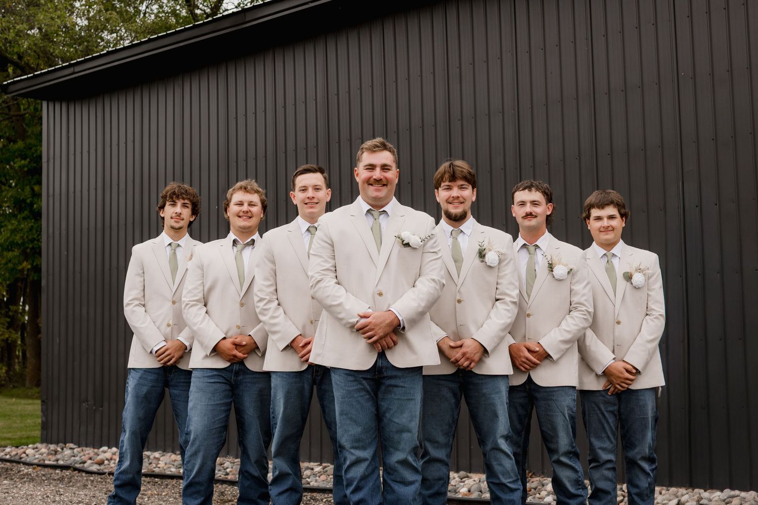 Groom and groomsmen in white blazers and jeans posing in front of a black barn at a rustic wedding.