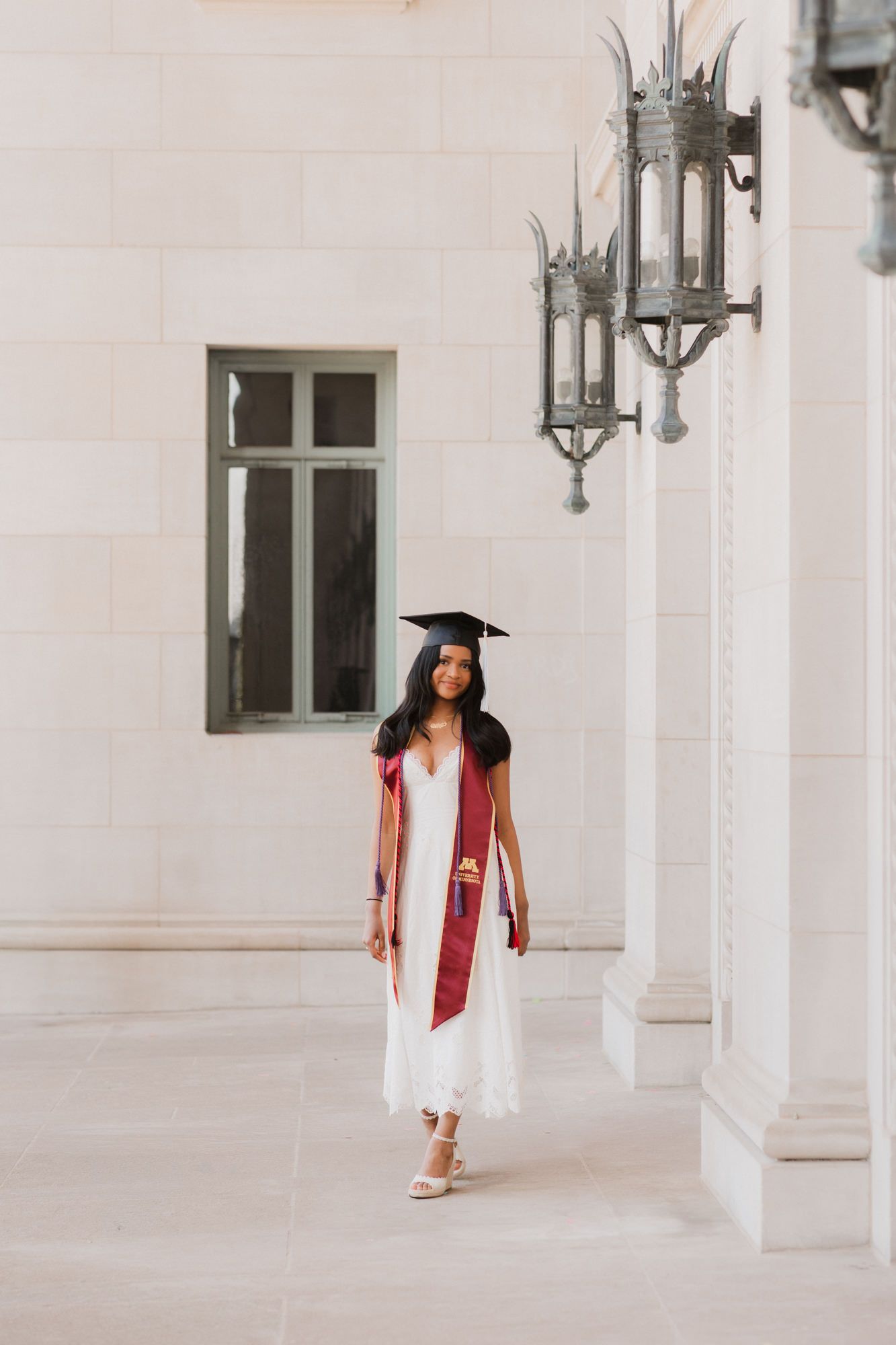 Young woman in white dress, graduation cap, and red sash poses between columns at a university building.
