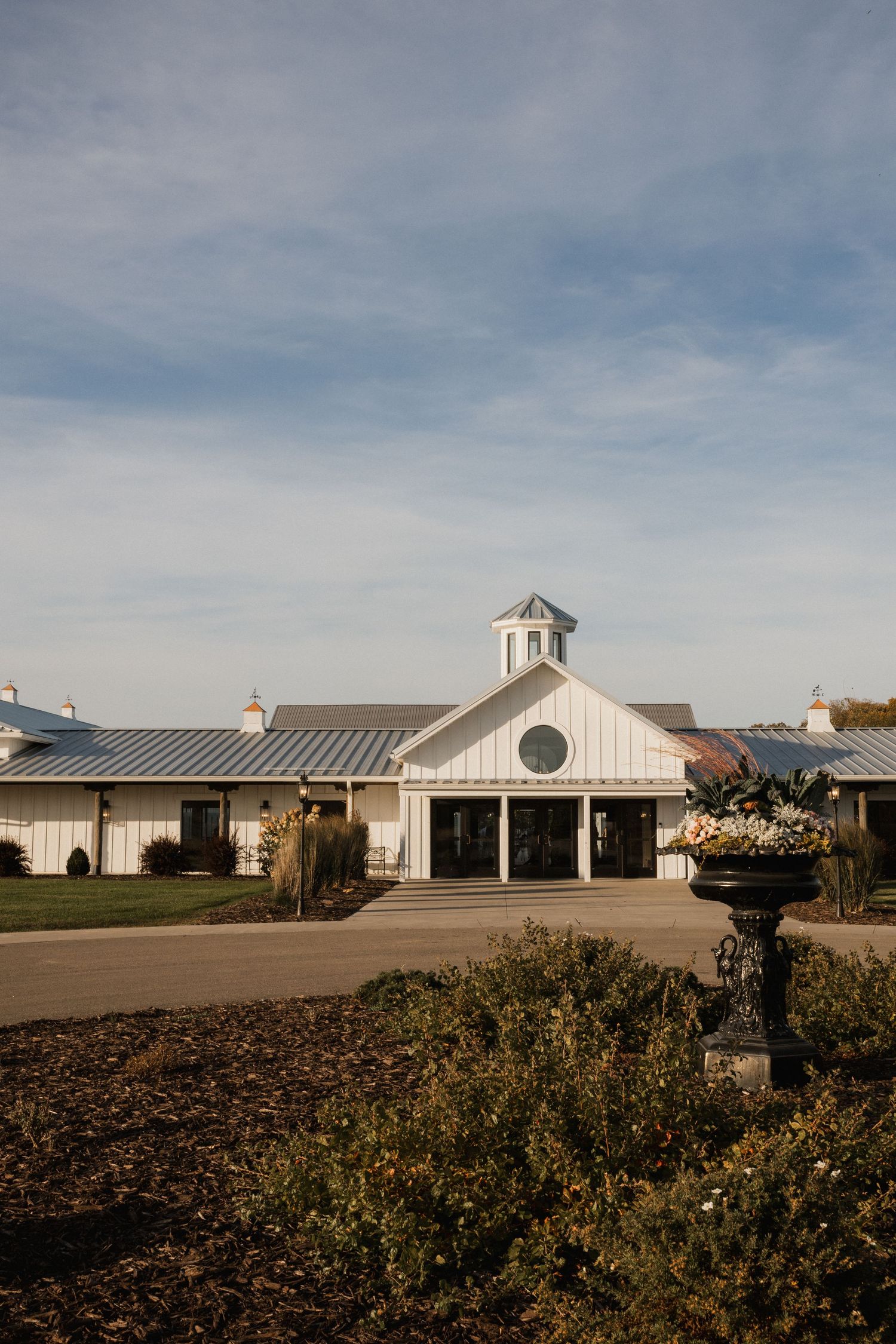 White barn-style wedding venue with a metal roof, cupola, and a planter of flowers in the foreground.