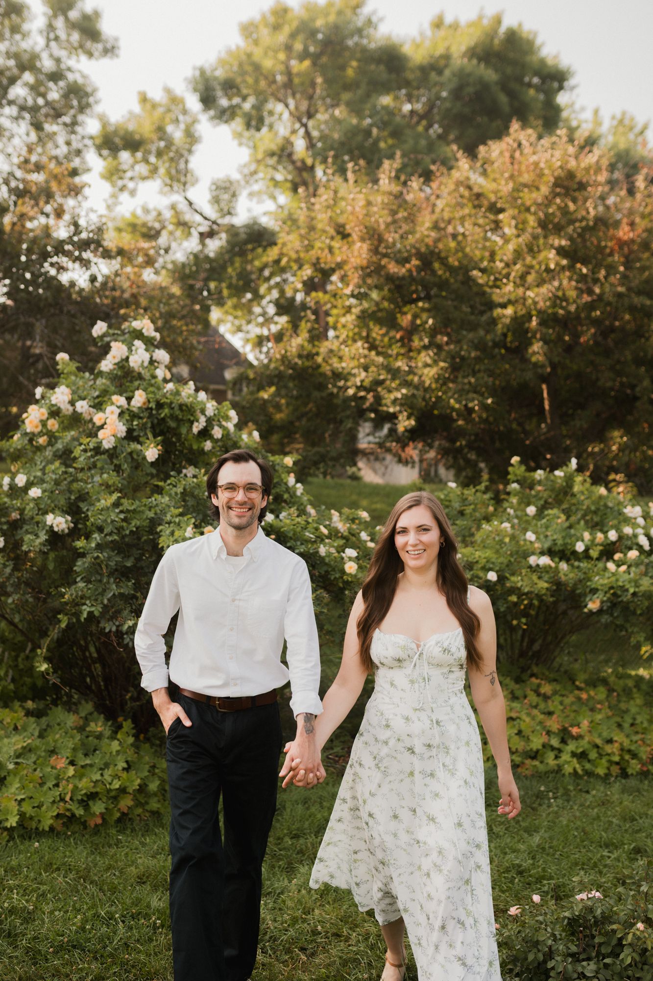 A smiling couple holds hands while walking through a lush garden with blooming roses and green trees in the background.