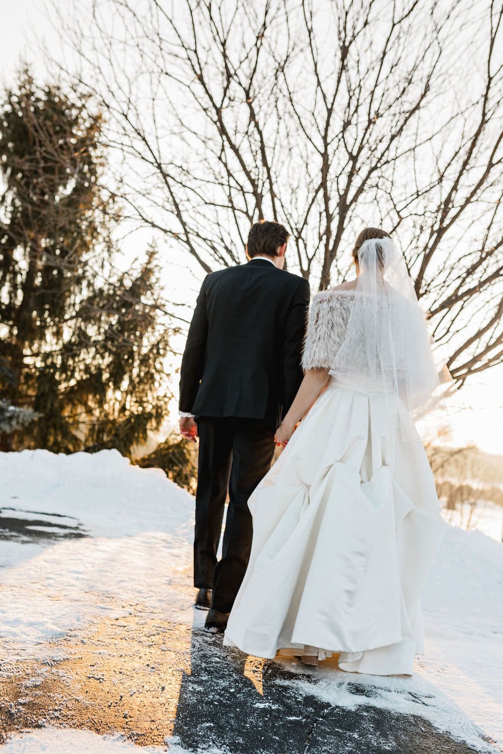 A wedding couple walks through a snowy landscape at sunset in formal attire.