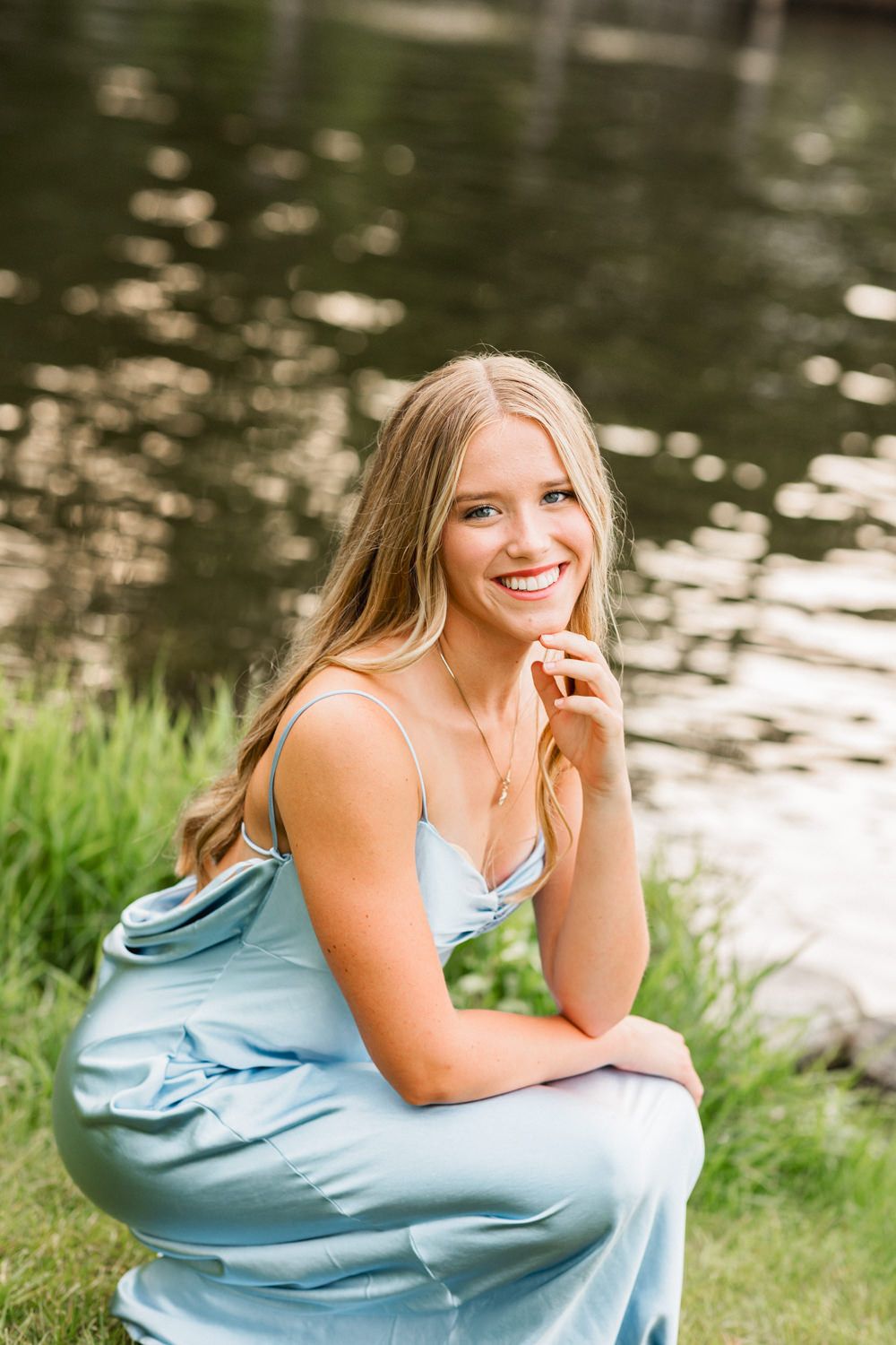 A light blue satin dress is worn by someone posing near a reflective pond on a sunny day.