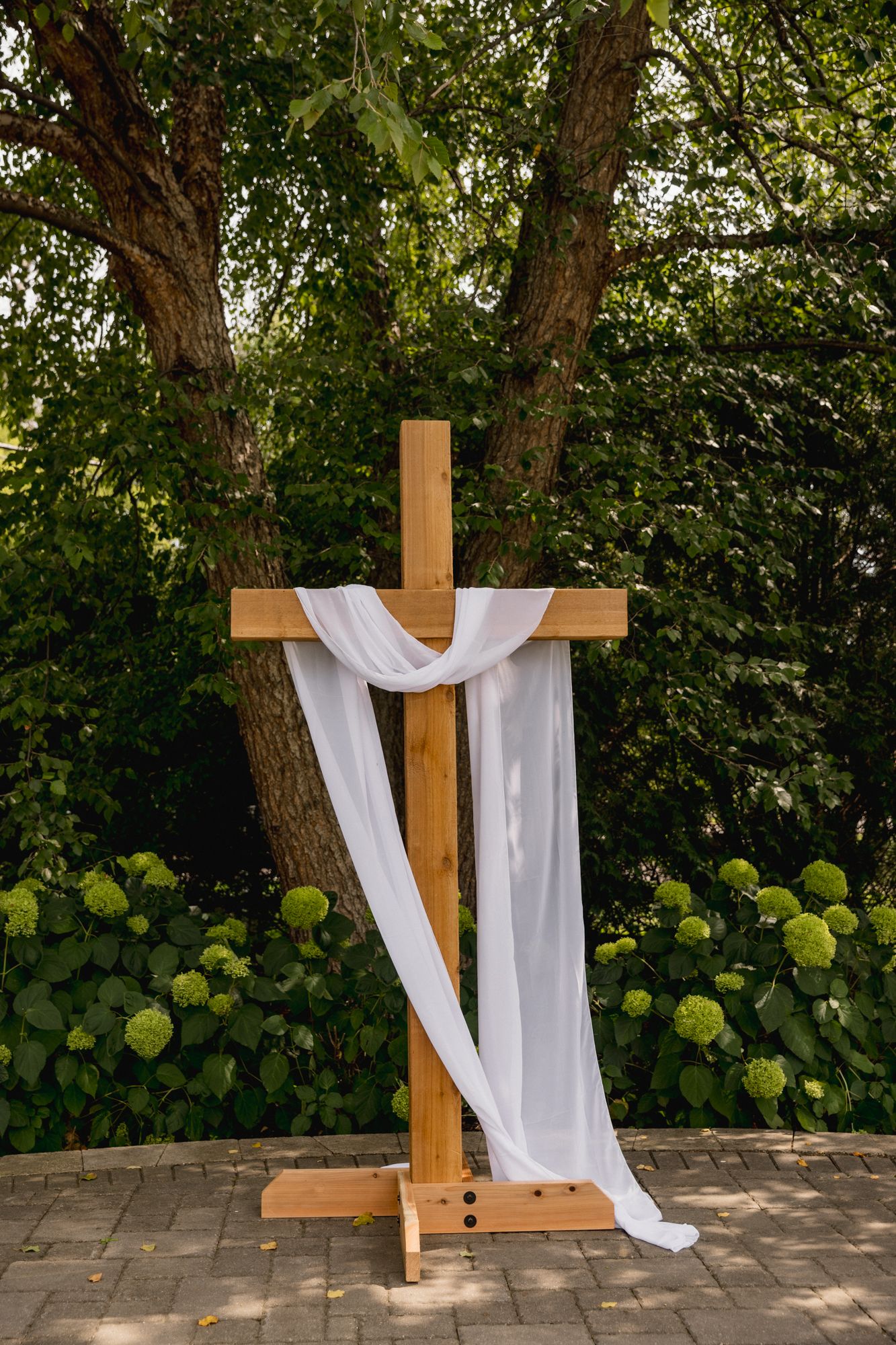 Wooden Christian cross draped with white fabric outdoors, surrounded by green trees and hydrangeas for a wedding ceremony.