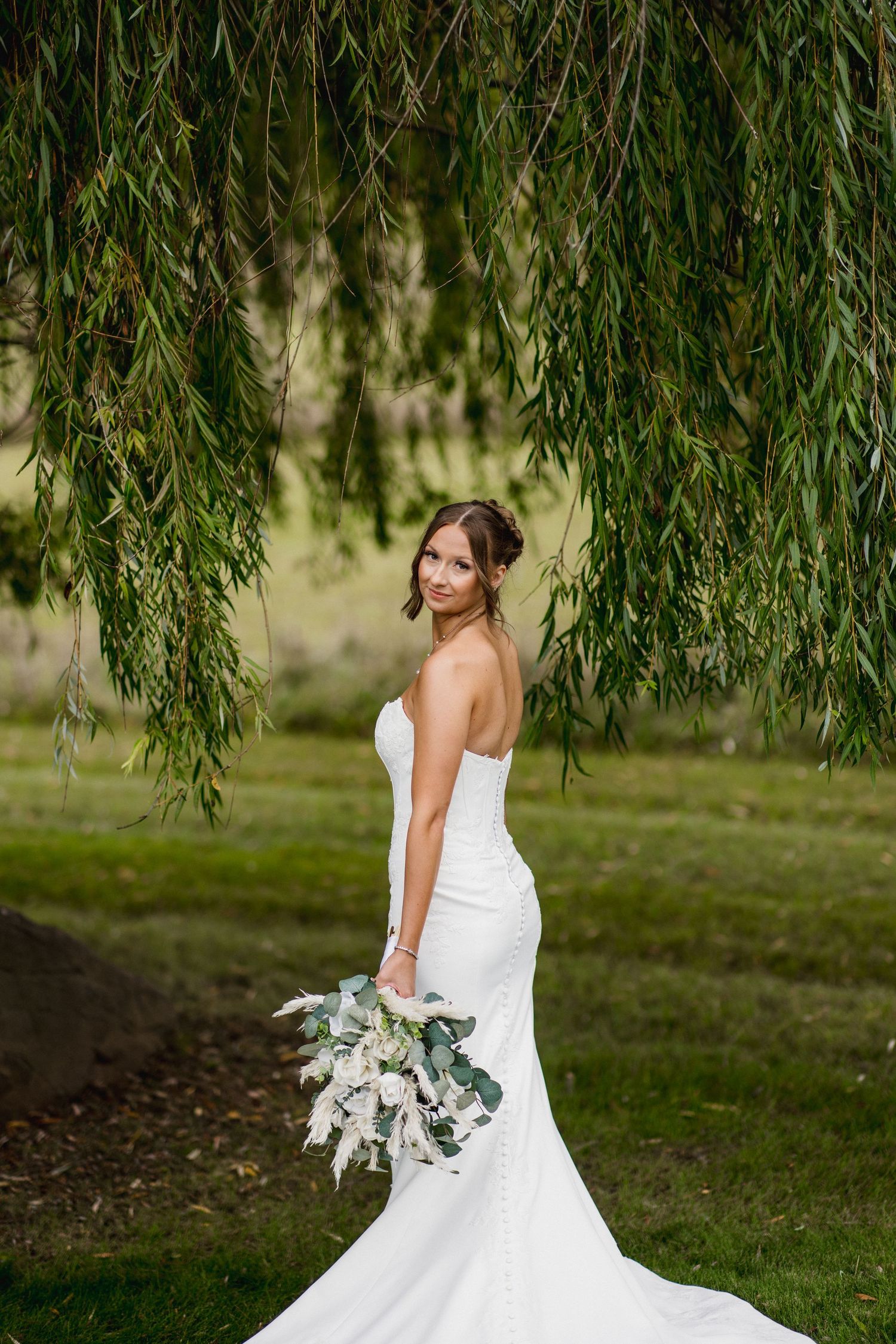 Bride in strapless white mermaid gown holds bouquet under weeping willow tree outdoors.