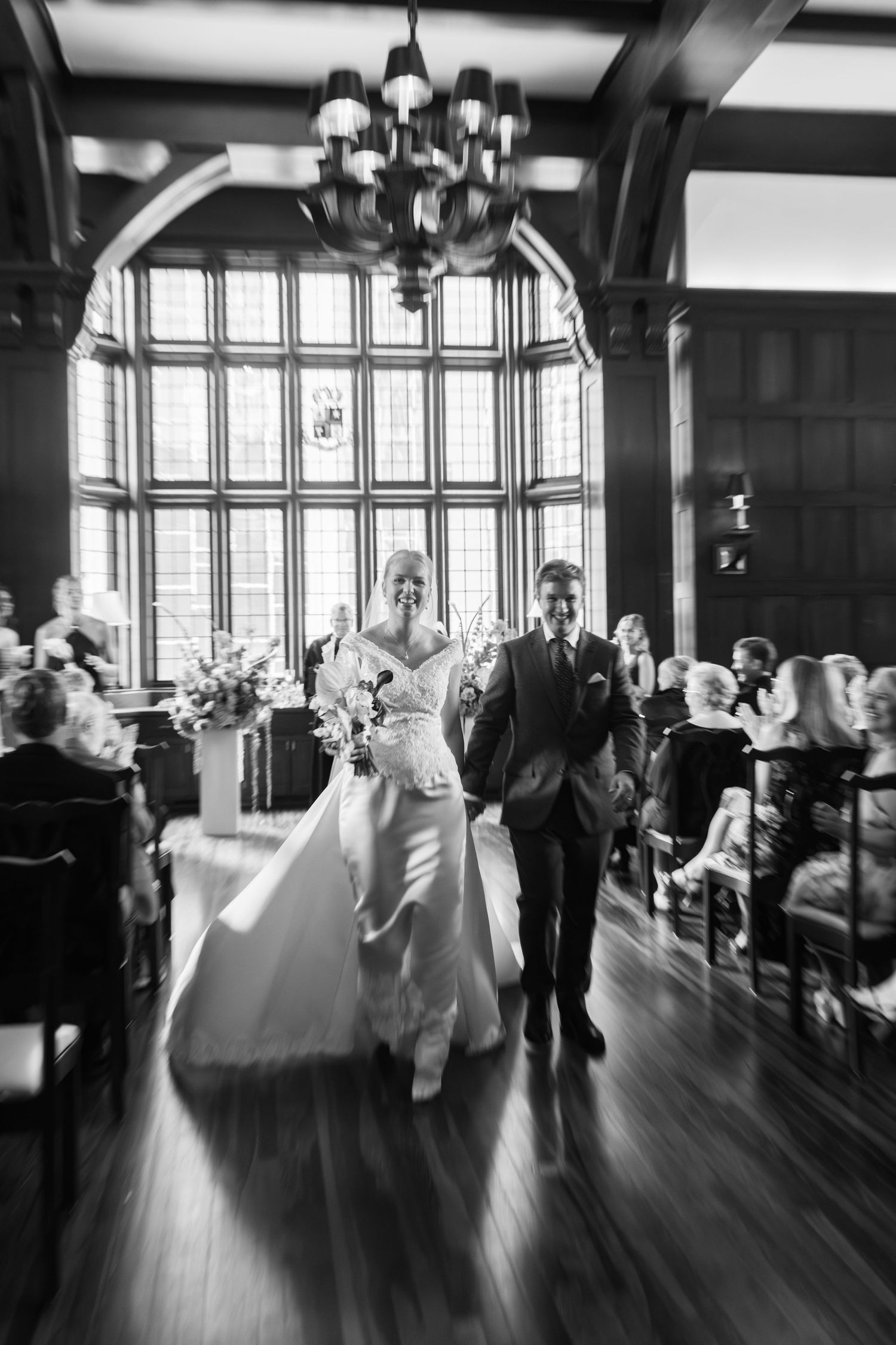 Black and white photo of bride and groom walking down the aisle after ceremony in an elegant wood-paneled hall.