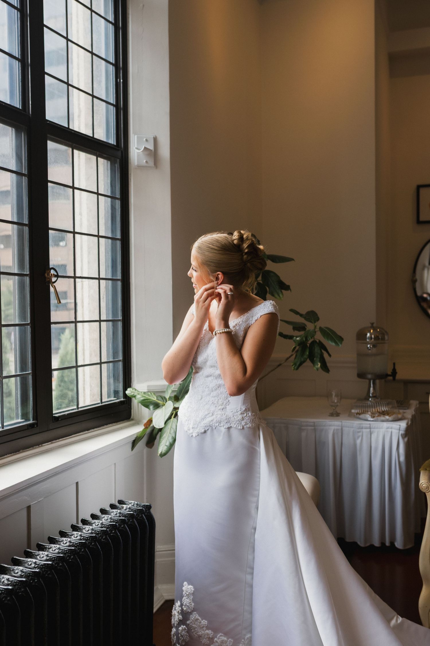Bride in lace wedding dress adjusts earring while standing by tall window in a softly lit room.