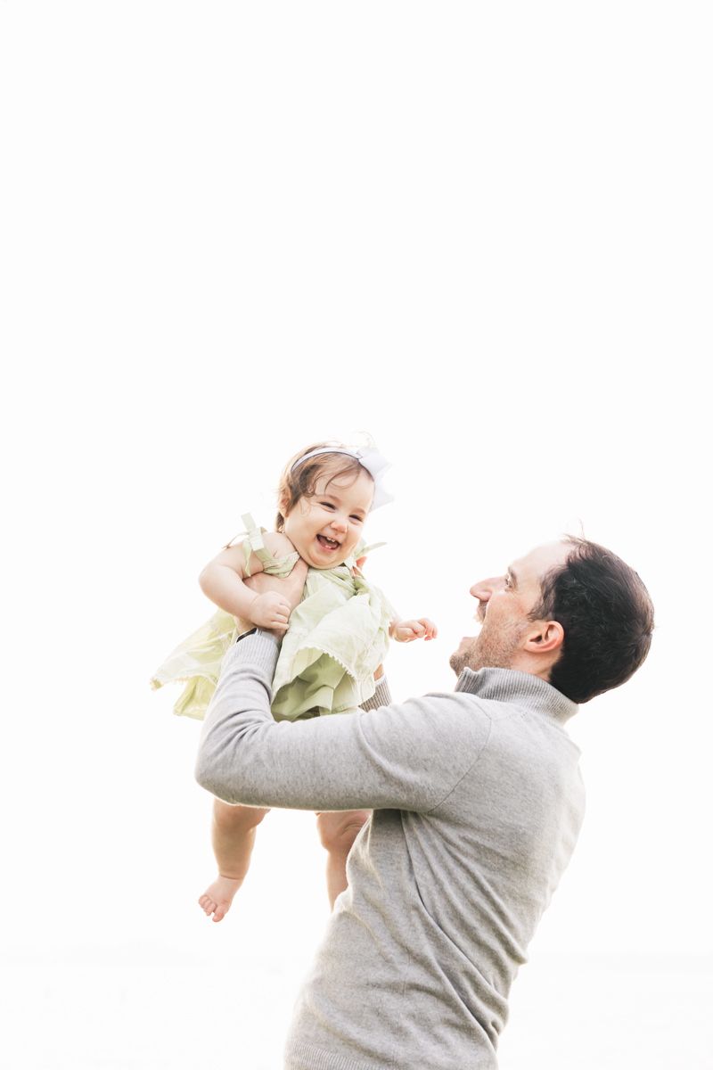 Parent lifts laughing child wearing yellow dress against white background.