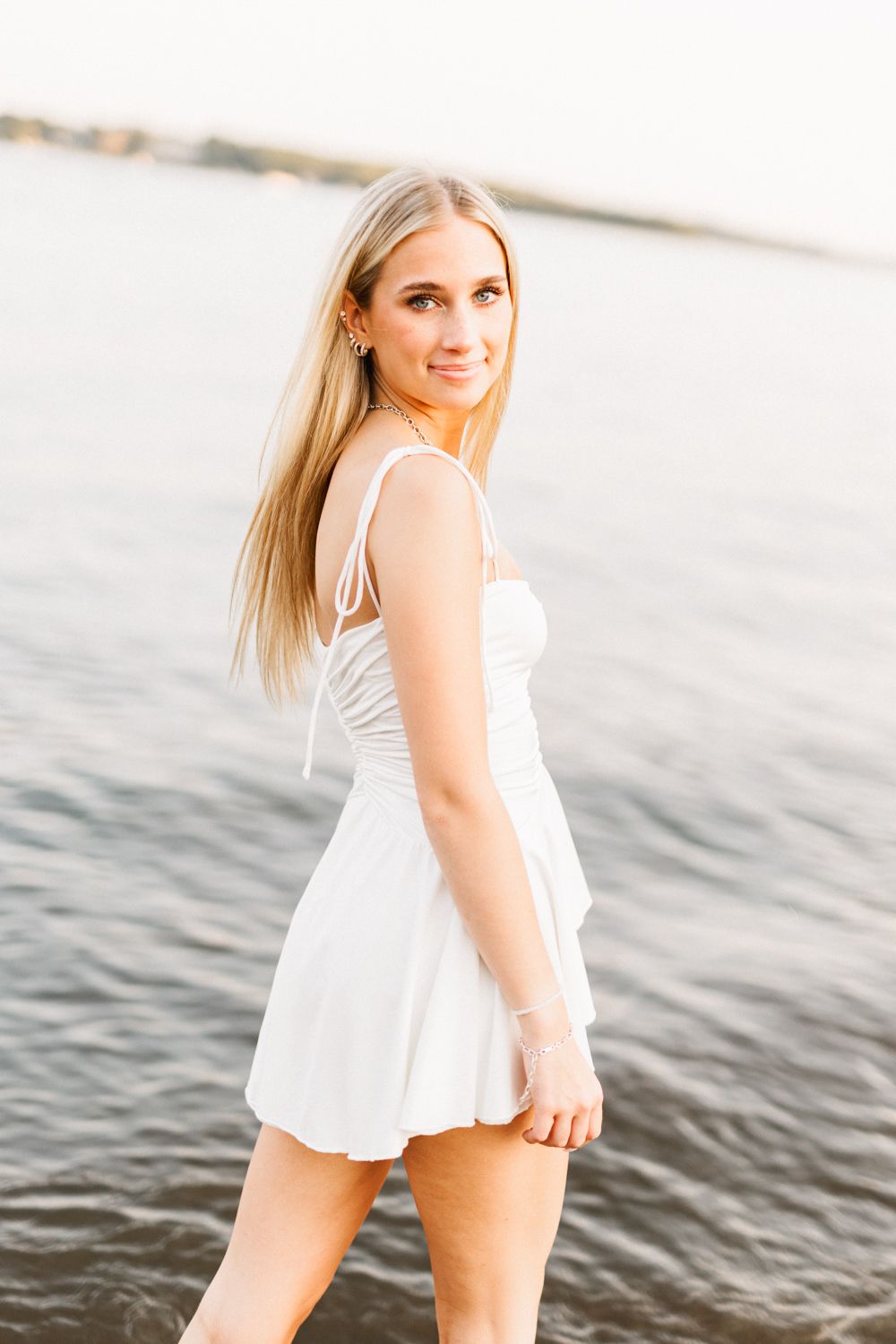 Woman in flowy white sundress poses near waterfront at golden hour.