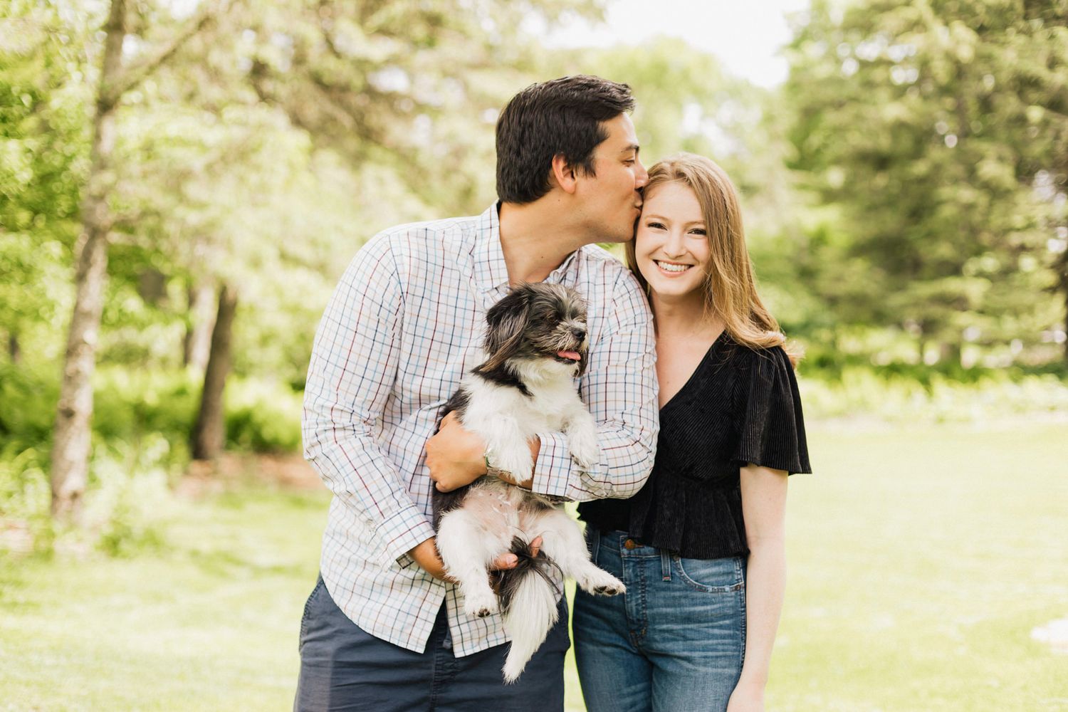 Couple holding small dog while sharing tender moment in sunny park setting.
