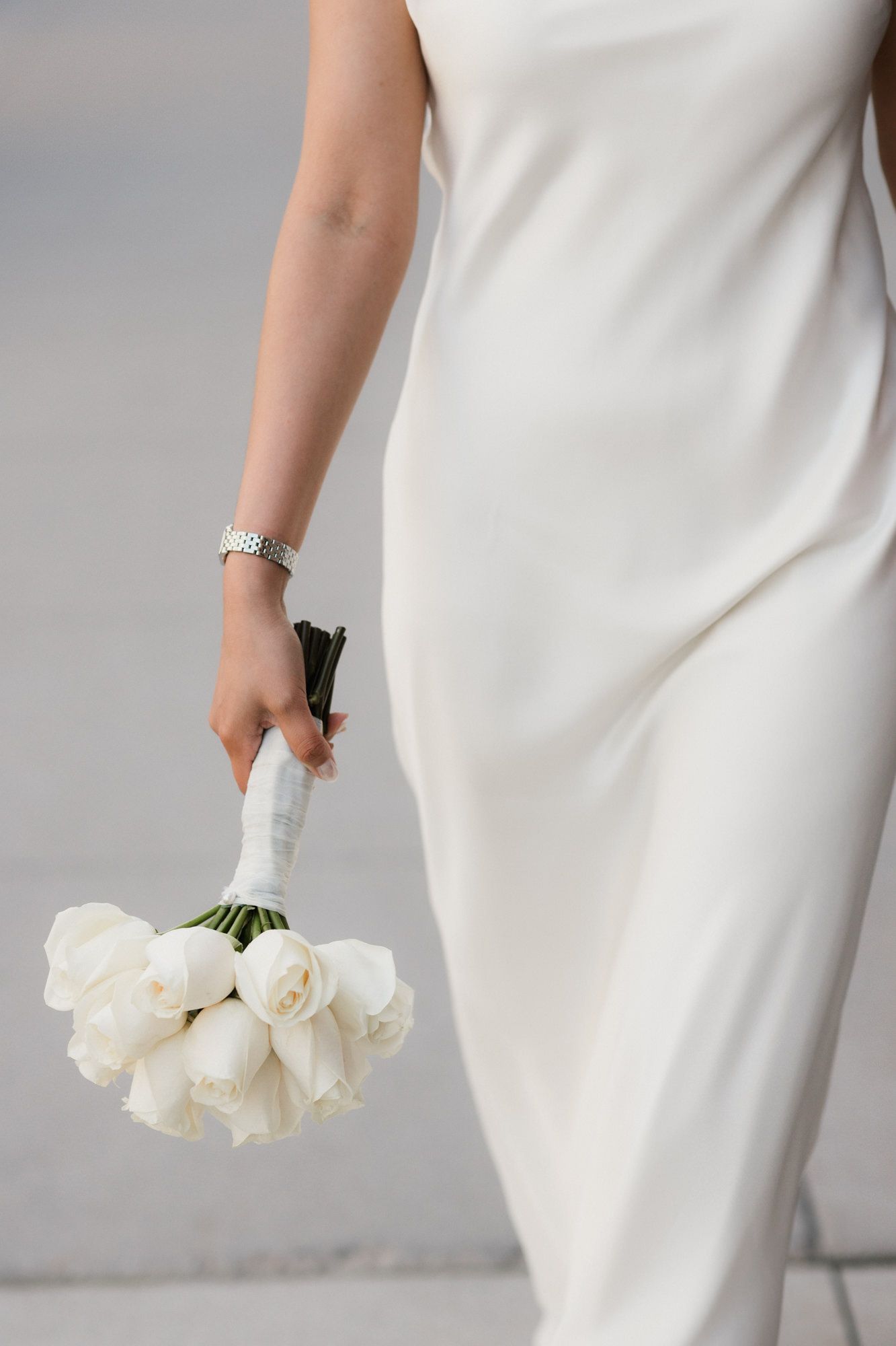 Bride in white satin dress holding elegant bouquet of white tulips tied with ribbon, wearing silver bracelet.