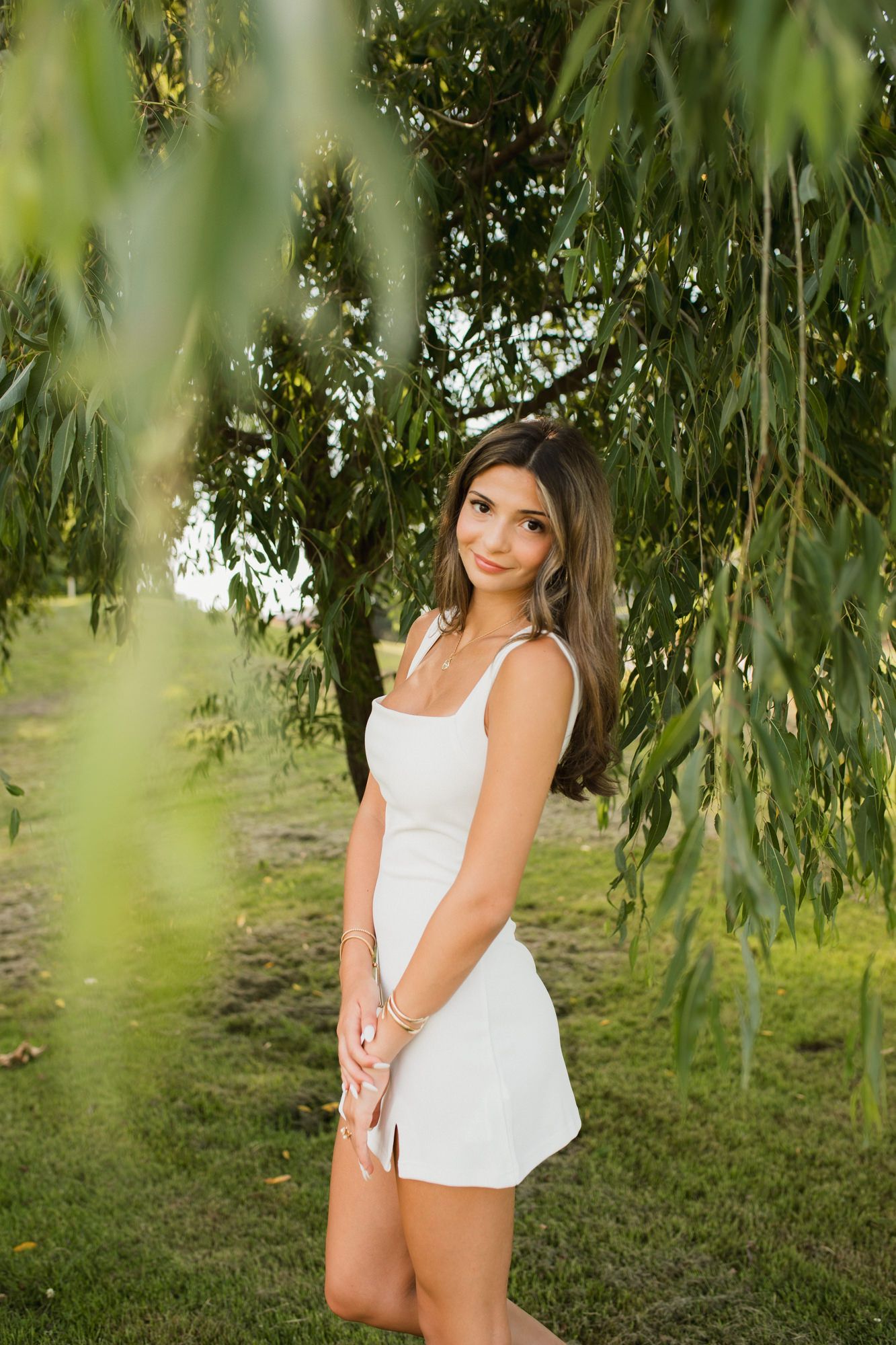 Young woman in white mini dress poses gracefully under weeping willow tree branches in lush green outdoor setting.