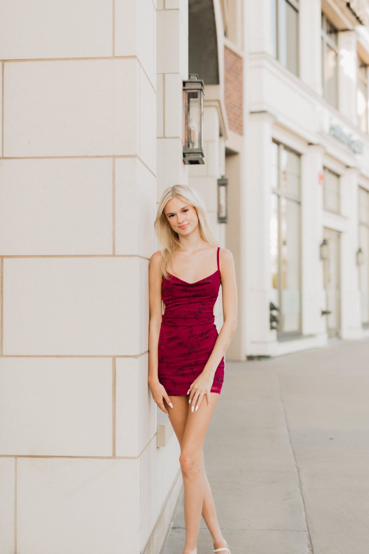 Blonde woman in a red mini dress leaning against a white building wall on a city sidewalk.