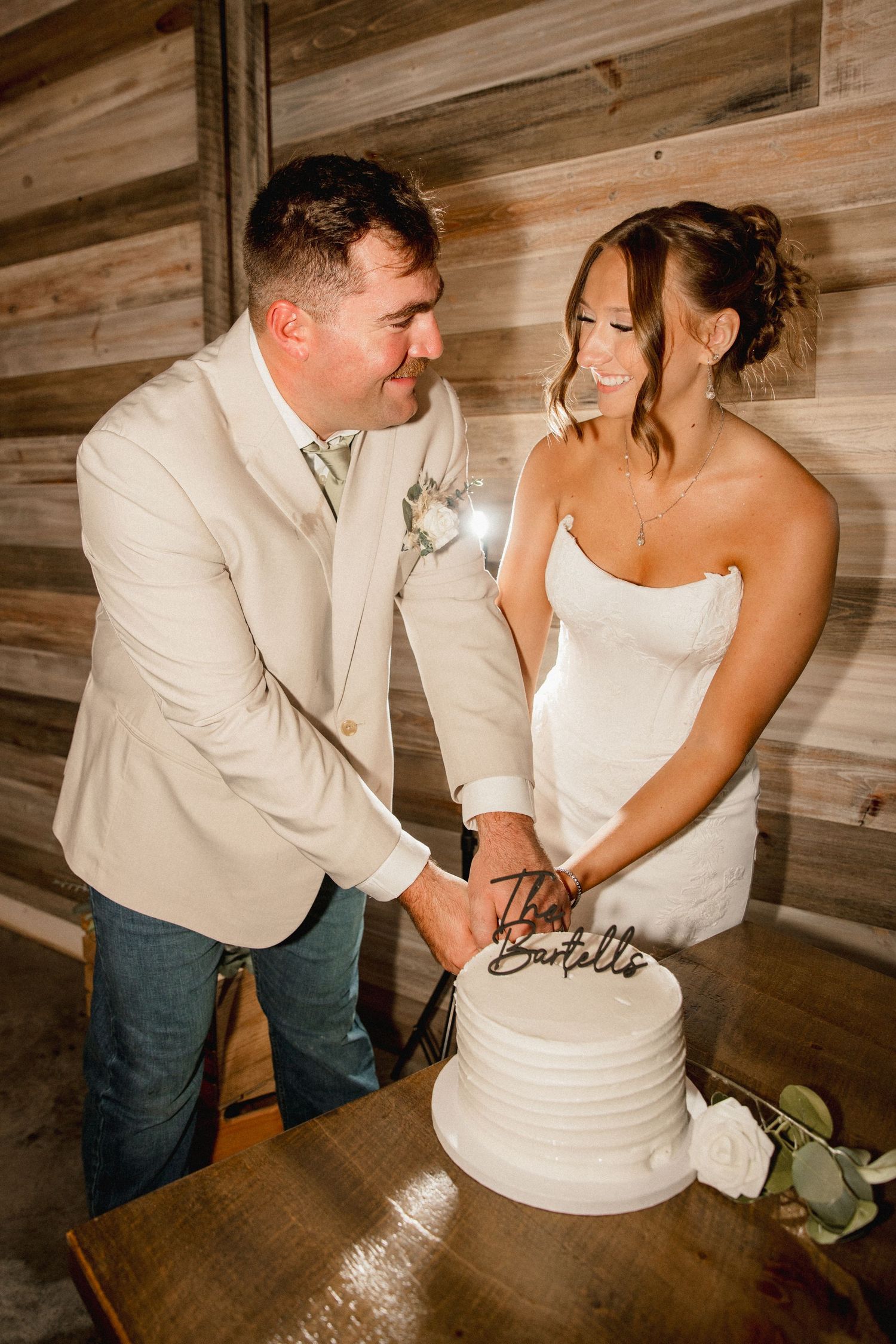 Bride and groom smiling while cutting their white tiered wedding cake together in a rustic barn venue.