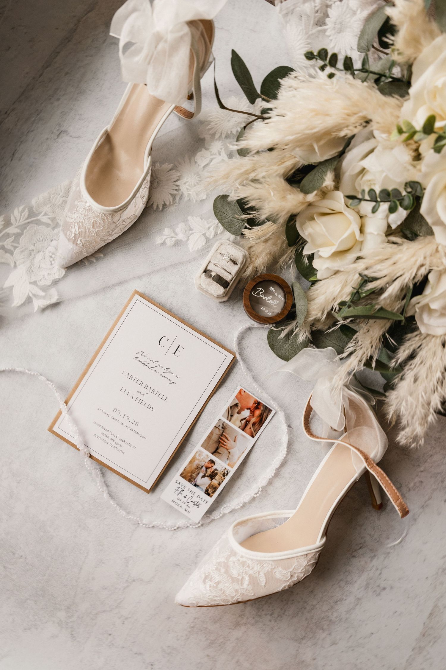 Elegant wedding flat lay with lace heels, floral bouquet, invitation suite, ring box, and pampas grass on marble.