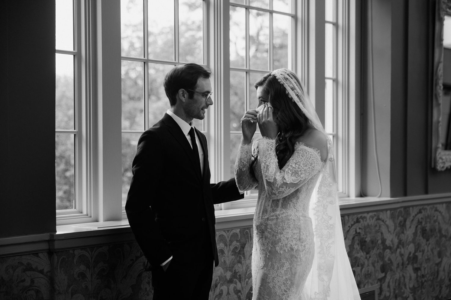 Black and white photo of groom holding brides hands near windows during emotional wedding first look moment.
