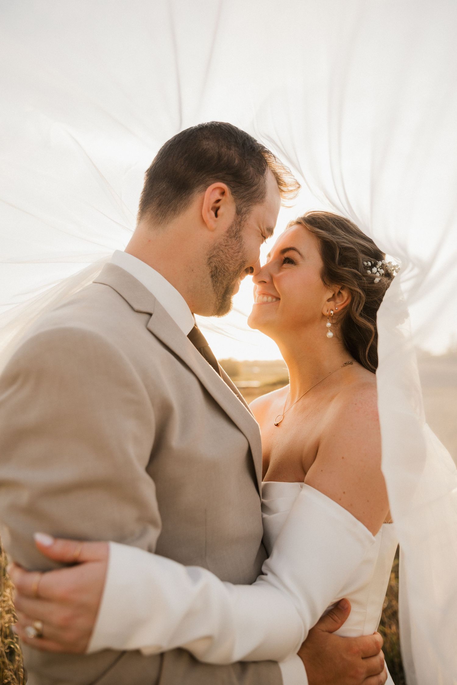 Smiling bride and groom share an intimate moment nose to nose as the bride's veil floats in the golden sunlight.